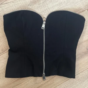 Svart korsett-topp från Zara XS - Supersnygg svart korsett-topp från Zara i storlek XS. Toppen är axelbandslös med en markerad dragkedja framtill som ger en edgy vibe. Stilren och tight passform, perfekt att styla med jeans eller kjol. Materialet känns mjukt och följsamt.