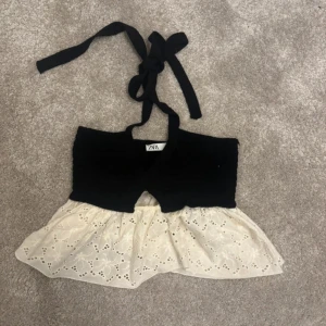 Svart och vit topp från Zara - Supersöt topp från Zara med svart stickad överdel och knytband i nacken. Nederdelen är vit med broderade detaljer och volang, vilket ger en romantisk vibe. Perfekt för varma dagar när du vill sticka ut lite extra.