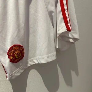 Manchester United vit fotbollsshorts - Vita shorts med röda ränder och Manchester Uniteds klubbmärke broderat på framsidan. Klassisk passform och tillverkad i lätt och bekvämt material, perfekt för dig som vill visa vilket lag du håller på.