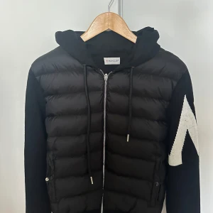 Svart Moncler pufferjacka med huva - Svart pufferjacka från Moncler med huva och dragkedja framtill. Jackan har stickade ärmar med vit zigzag-detalj och Moncler-logga på ena ärmen. Kroppen är quiltad och vadderad, perfekt för kyliga dagar. Snygg och sportig look med coola kontraster.