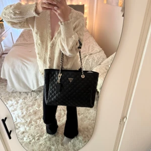 Svart quiltad axelväska från Guess - Säljer en svart quiltad axelväska från Guess med silverfärgade kedjedetaljer och logga framtill. Väskan har rejäla handtag i skinn och kedja, samt rymligt beige innerfoder med flera fack och dragkedja. Perfekt för dig som vill ha plats för allt du behöver.🖤🌟🥰