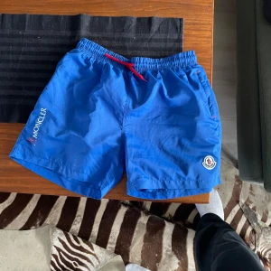 Blå Moncler badshorts med snörning - Snygga blå badshorts från Moncler med röd snörning i midjan och klassisk Moncler-l på benet. Shortsen har elastisk midja och bakficka med knapp. Dom är xxl men sitter som L skulle jag sägs, skannar man qr koden så ser man att dom ät rikiga. Nätet är klippt för att det skaver mycket.