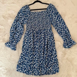 Blå blommig klänning med långärmad puffärm - Supersöt blå kortklänning med vita blommor och långa puffärmar med volang vid ärmslut. Fyrkantig uringning och flowig kjol