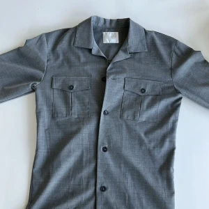 Grå overshirt från Oscar Jacobson S - Stilren grå overshirt från Oscar Jacobson i klassisk modell med två bröstfickor med lock och knappar framtill. Tunn och mjuk känsla, perfekt för en clean look. Passar dig som gillar minimalistisk stil och snygga detaljer.