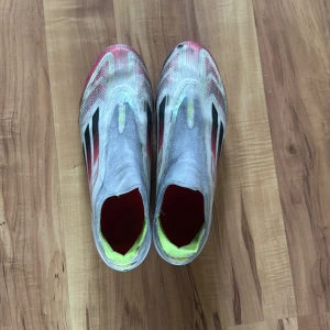 Adidas F50 fotbollsskor i vitt och rött - Säljer ett par Adidas F50 fotbollsskor i vitt med röda och svarta detaljer samt gula inslag. Skorna har en strömlinjeformad design utan klassisk snörning och är tillverkade i lätt syntetmaterial för maximal fart på planen. Har dock en liten skada i fram men det är inga större problem 