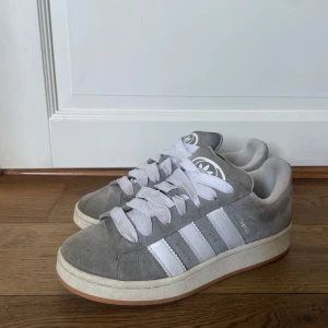 Grå Adidas Campus sneakers i mocka - Snygga Adidas Campus sneakers i grå mocka med klassiska vita ränder och breda vita skosnören. Låg modell med rund tå och vit sula. Perfekta för dig som gillar en clean och tidlös streetstil.