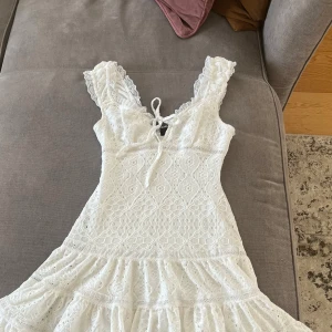 Vit populär spetsklänning med volang - Superfin vit kortklänning i spets med volangig kjol och v-ringning med snörning.Perfekt för sommarens alla tillfällen!Den har en sminkfläck på insidan men inget man tänker på alls.Endast använd en gång!