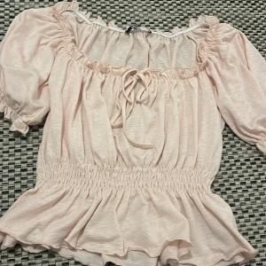 Ljusrosa blus från Gina Tricot - Supersöt ljusrosa blus från Gina Tricot med puffiga korta ärmar, smockad midja och volangkant nertill. Blusen har knytning framtill och en lätt rynkad struktur i tyget. Perfekt för dig som gillar romantisk och trendig stil.