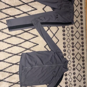 Grå träningsset från Under Armour - Säljer ett grått träningsset från Under Armour med svarta detaljer längs sidorna. Setet består av en zip-jacka och matchande byxor, perfekt för dig som gillar sportig och stilren look. Materialet är lätt och smidigt, passar bra till träning eller uppvärmning.