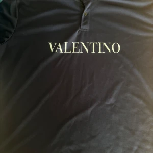 Mörkblå pikétröja från Valentino - Snygg mörkblå pikétröja från Valentino med klassisk krage och knäppning framtill. Stor vit logga tryckt över bröstet ger en clean och trendig look. Perfekt till jeans eller shorts för en stilren vibe.