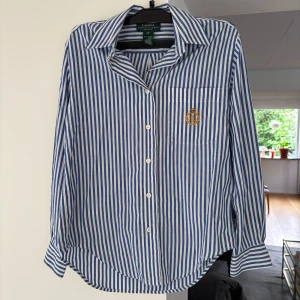 Randig skjorta från Ralph Lauren - Snygg blåvit randig skjorta från Ralph Lauren med klassisk krage och knäppning framtill. Bröstficka med broderad RL-logo i guld och krona. Tillverkad i 100% bomull.