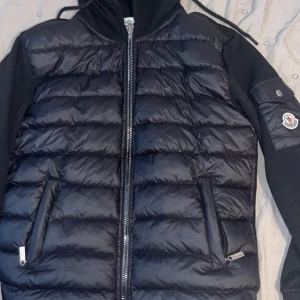 Svart pufferjacka från Moncler - Snygg svart pufferjacka från Moncler med quiltad framsida och ärmar i mjuk bomull. Jackan har huva med dragsko, dragkedja framtill och två fickor med dragkedja. Moncler-logga på ena ärmen ger en exklusiv vibe.