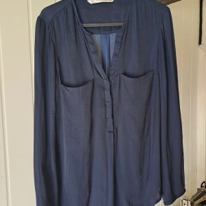 Mörkblå Zara blus  - Mörkblå blus från Zara Trafaluc med lång ärm och djup v-ringning. Modellen är enkel och flowy med två stora fickor framtill. Perfekt för dig som gillar stilrena plagg med en chill vibe.