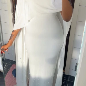 Vit långklänning med cape - Säljer en elegant vit långklänning med cape-detalj. Klänningen har figurnära passform och svepande tyg som ger en dramatisk look. Perfekt för dig som vill sticka ut med en stilren och modern känsla. Materialet känns mjukt och följsamt mot kroppen.