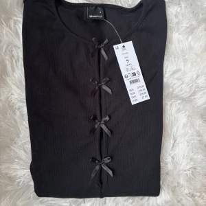 Svart långärmad topp med rosetter Gina Tricot - Supersnygg svart långärmad topp från Gina Tricot med ribbad struktur och fyra söta rosetter framtill. Rund halsringning och figurnära passform. Perfekt att styla med jeans eller kjol för en trendig look.