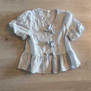 Randig blus med knyt från Gina Tricot - Supersöt blus från Gina Tricot i ljusblått och vitt med tunna ränder. Puffiga korta ärmar, v-ringning och tre knyt framtill. Volang nertill ger en extra touch. Perfekt för dig som gillar en romantisk och trendig stil.