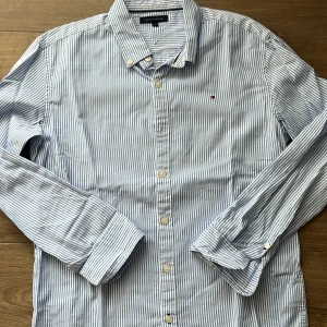 Randig skjorta från Tommy Hilfiger - Snygg skjorta från Tommy Hilfiger i vitt och ljusblått med tunna vertikala ränder. Klassisk krage, knäppning framtill och broderad logga på bröstet. Tillverkad i mjuk bomull och har långa ärmar med manschettknappar. Perfekt för en clean och stilren look.
