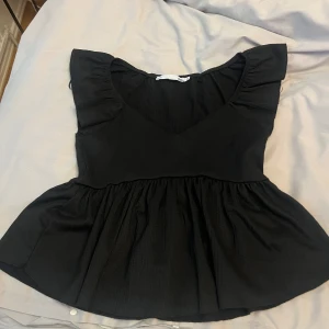 Svart blus med volang från 157 - Supersöt svart blus från 157 med v-ringning och volangdetaljer vid axlarna. Toppen har en lätt rynkad, utställd nederdel och är tillverkad i ett mjukt, strukturerat tyg. Perfekt för dig som gillar en feminin och trendig look.