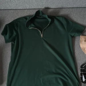 Mörkgrön pikétröja  - Snygg mörkgrön pikétröja från DELE i storlek S. Tröjan har korta ärmar, klassisk krage och en trendig half zip-dragkedja framtill. Perfekt för dig som gillar stilrena och sportiga vibes. Materialet känns mjukt och bekvämt mot huden.
