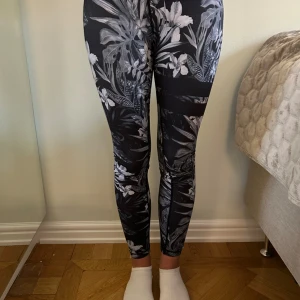 Stronger leggings med blommigt mönster - Leggings från Stronger med grått och vitt blommigt mönster. Hög midja med logga i midjebandet och tight passform. Tillverkade i stretchigt syntetmaterial. 