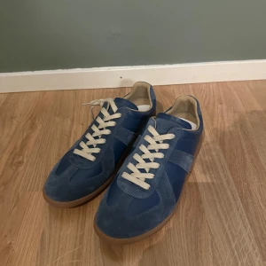 Blå sneakers från Maison Margiela - Snygga blå sneakers från Maison Margiela med ovandel i mocka och skinn, beige snörning och klassisk gummisula. Skorna har en stilren design med rund tå och diskreta detaljer, perfekta för dig som gillar en clean look.