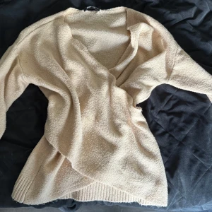 Beige omlottstickad kofta - Mysig beige stickad kofta med omlott-design och v-ringning. Tröjan har långa ärmar och ribbade muddar vid ärmslut och nederkant. Perfekt för lager-på-lager och chill dagar.