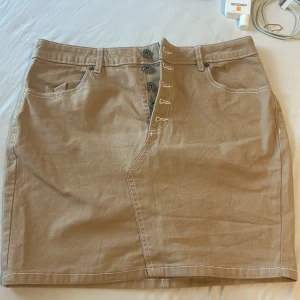 Beige jeanskjol med knappar - Snygg beige kort jeanskjol med markerade vita sömmar och fyra knappar framtill. Kjolen har klassiska fickor både fram och bak samt hällor för bälte. Perfekt för dig som gillar en enkel men trendig look.