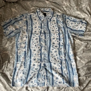 Blåvit kortärmad hawaiiskjorta  - Snygg skjorta i blått och vitt med blommigt mönster och korta ärmar. Perfekt för sommaren med avslappnad passform och knappar framtill. Skjortan har klassisk krage och tropisk vibe, perfekt för dig som vill sticka ut lite extra. Säljer mycket andra skjortor så köp gärna flera i ett bundle!