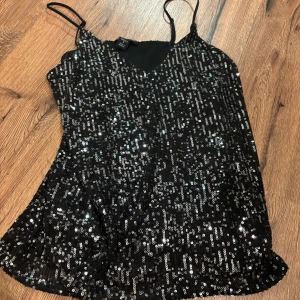 Svart glitter topp - Glittrig svart topp täckt av paljetter med smala axelband och v-ringning. Perfekt för dig som vill sticka ut på festen! Snyggt flowy fall och riktigt partyvibe.