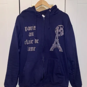 Mörkblå hoodie med strass och tryck - Mörkblå hoodie från BLINGLUCID med dragkedja, huva och fickor. Framsidan har strasstenar som bildar texten 'paris au clair de lune' och motiv av Eiffeltornet och måne. Tillverkad i bomull och polyester, perfekt för dig som gillar statement-plagg. Finns inga tecken på slitage eller användning. Säljs pågrund av att den är för liten. Kan släppas billigare 