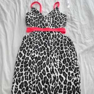 Leopardmönstrad klänning med neonrosa detaljer - Snygg kortklänning med svart och grått leopardmönster på vit botten. Klänningen har smala neonrosa axelband och ett matchande neonrosa band i midjan. Ärmlös modell med cut-out vid midjan för en trendig look. Perfekt för dig som vill sticka ut.