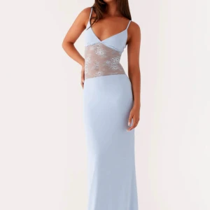 Ljusblå långklänning med spetsdetalj - Supersnygg ljusblå långklänning med smala axelband och v-ringning. Klänningen har en transparent spetsdetalj i midjan och öppen rygg, vilket ger en extra cool och trendig look. Perfekt för dig som vill sticka ut med en elegant men modern stil.