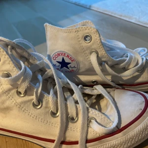 Vita Converse Chuck Taylor All Star - Klassiska vita Converse Chuck Taylor All Star high-tops med röd och blå rand längs sulan. Skorna har snörning, rund tå och är tillverkade i canvas. Perfekta för en avslappnad och cool streetstil.