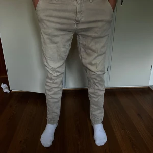 Beige Replay Hyperflex chinos - Snygga beige chinos från Replay Hyperflex-serien, modell Zeumar. Byxorna har klassisk passform med raka ben, bälteshällor och bakfickor med knapp. Stretchigt material för extra komfort och stilren look.