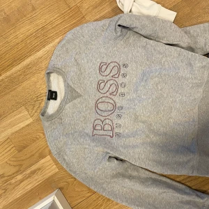 Grå sweatshirt från Hugo Boss - Säljer en stilren grå sweatshirt från Hugo Boss med röd text på bröstet. Tröjan har långa ärmar och en rund halsringning. Perfekt för en avslappnad stil.