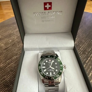 Swiss Alpine Military klocka - Säljer en stilren Swiss Alpine Military klocka med en grön och svart vridbar bezel. Klockan har en svart urtavla med vita markeringar och en robust länkarmband i silverfärgat stål. Perfekt för den som gillar en sportig och elegant stil.