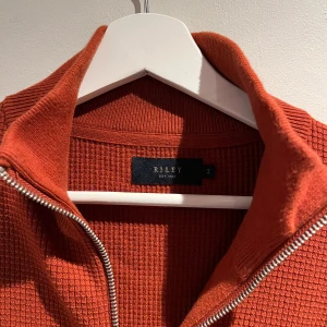 Rostfärgad ribbad tröja från Riley - Säljer en snygg rostfärgad ribbad tröja från Riley med dragkedja framtill. Perfekt för lager-på-lager och har en stilren design med långa ärmar. Passar bra till både jeans och chinos.