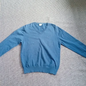 Blå v-ringad tröja - Säljer en stilren blå v-ringad tröja med ett litet broderat märke på bröstet. Tröjan har långa ärmar och ribbade kanter vid ärmslut och nederkant. Perfekt för en avslappnad look.