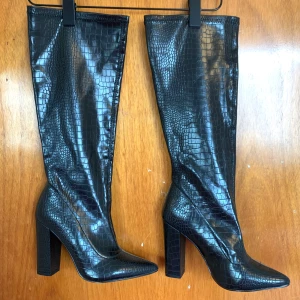 Svarta stövlar med krokodil/orm mönster - Snygga svarta stövlar med krokodilmönster och hög klack. De har en spetsig tå och är perfekta för att ge en elegant touch till din outfit. Tillverkade i syntetmaterial.