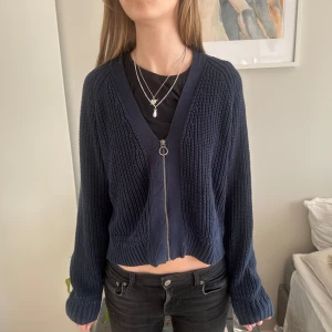 Mörkblå stickad kofta med dragkedja - Säljer en snygg mörkblå stickad kofta med dragkedja framtill. Koftan har en v-ringad design och långa ärmar, perfekt för lager-på-lager-stil. Den ribbade strukturen ger en extra touch av stil. Passar perfekt till både jeans och kjol.