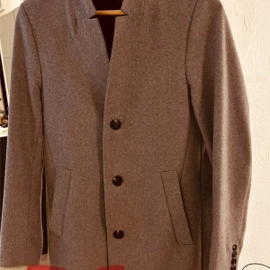 Grå kappa med knappar - Elegant grå kappa med stilrena knappar framtill. Den har en klassisk design med långa ärmar och två fickor på sidorna. Perfekt för en stilren look.