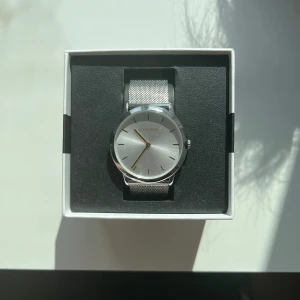 Silverfärgad klocka från Calvin Klein - Elegant klocka från Calvin Klein med en stilren silverfärgad urtavla och matchande mesh-armband. Klockan har en minimalistisk design med subtila markeringar och en smal boett. Perfekt för den som gillar en tidlös och sofistikerad stil.