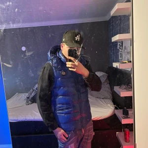 Blå dunväst från Bvaldi - Snygg blå dunväst från Bvaldi med dragkedja och två fickor framtill. Perfekt för lager-på-lager under kyliga dagar. Västen har en stilren design med ett litet märke på bröstet. Man kan även ta av luvan. Använd 2-3 gånger