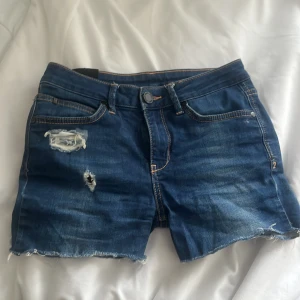 Blå jeansshorts  - Säljer ett par snygga blå jeansshorts med slitna detaljer och fransig kant. Perfekta för en avslappnad stil. De har klassisk femficksdesign och knappgylf.