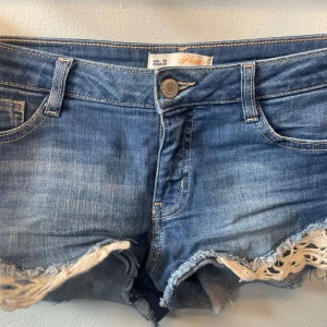 Blå jeansshorts från Clockhouse - Snygga blå lågmidjade jeansshorts från Clockhouse med fransiga kanter och spetsdetaljer. 