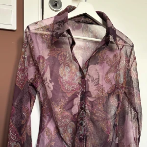 Mesh mönstrad skjorta / blus - Säljer en unik och snygg blus skjorta med ett paisleymönster i lila och orangea nyanser. Blusen har långa ärmar, vid ärmsluten finns rosett detaljer. Perfekt för en unik och stilfull look. Köpt på utvald vintage butik. Fairycore, witchcore. 