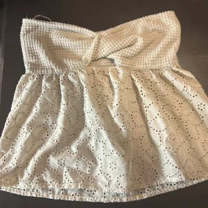 Vit axelbandslös topp med spetsdetaljer - Säljer en söt vit axelbandslös topp med elastisk överdel och vacker spetsdetalj nedtill. Perfekt för sommarens varma dagar. Toppen har en smockad överdel som ger en bekväm passform. 