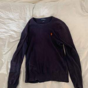 Säljer en stilren mörkblå tröja från Ralph Lauren med det klassiska broderade logotypen i orange på bröstet. Tröjan har långa ärmar och en rund halsringning. Perfekt för en avslappnad men ändå elegant look. Lappen vid nacken har släppt, bara ta bort eller sy fast igen. Inget man stör sig på.