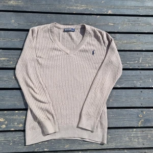Beige stickad tröja från Polo Ralph Lauren - Säljer en stilren beige stickad tröja från Polo Ralph Lauren med v-ringning och det klassiska logotypbroderiet på bröstet. Tröjan har långa ärmar och en ribbad struktur som ger en snygg look. Perfekt för kyligare dagar!