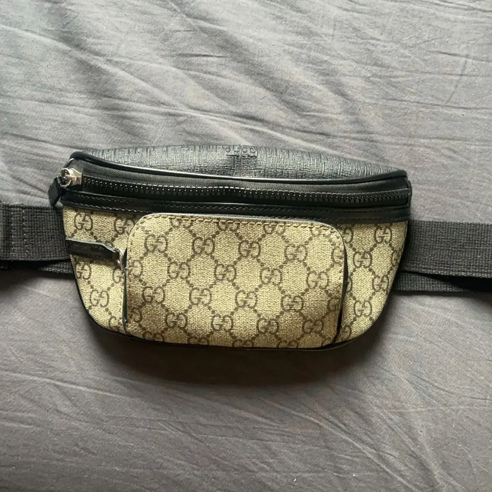 Snygg midjeväska från Gucci med det klassiska GG-mönstret i beige och svart. Väskan har flera dragkedjeförsedda fack och en justerbar rem för bekväm passform. Perfekt för att hålla dina saker säkra och stiliga. Väskan är äkta . Laukut & Käsilaukut.